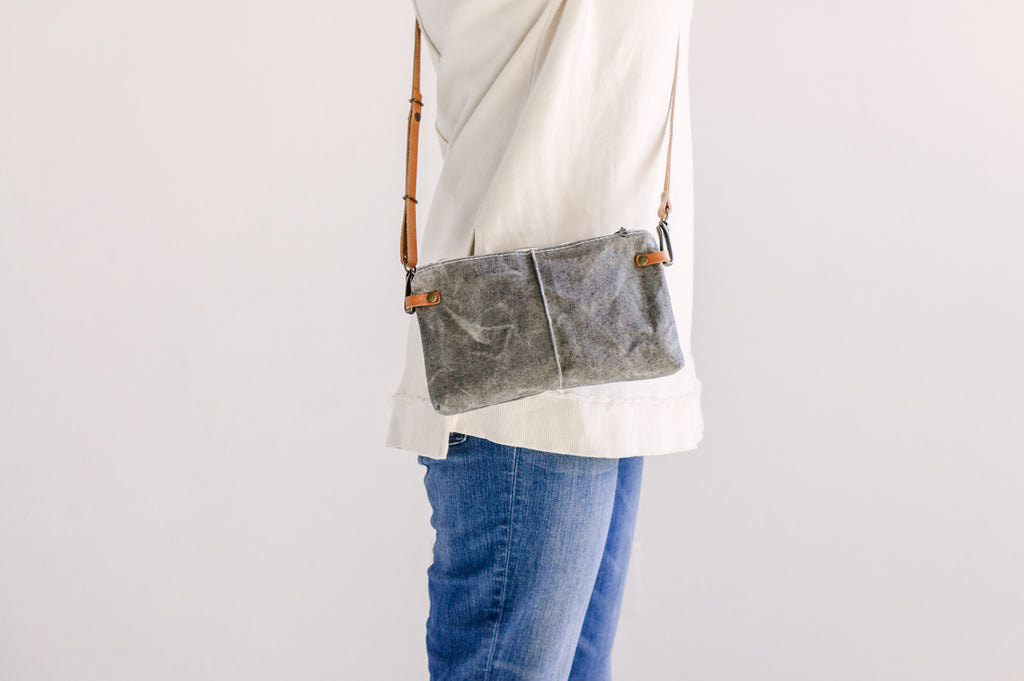 a gray waxed denim bag is worn as a crossbody clutch with brown leather straps