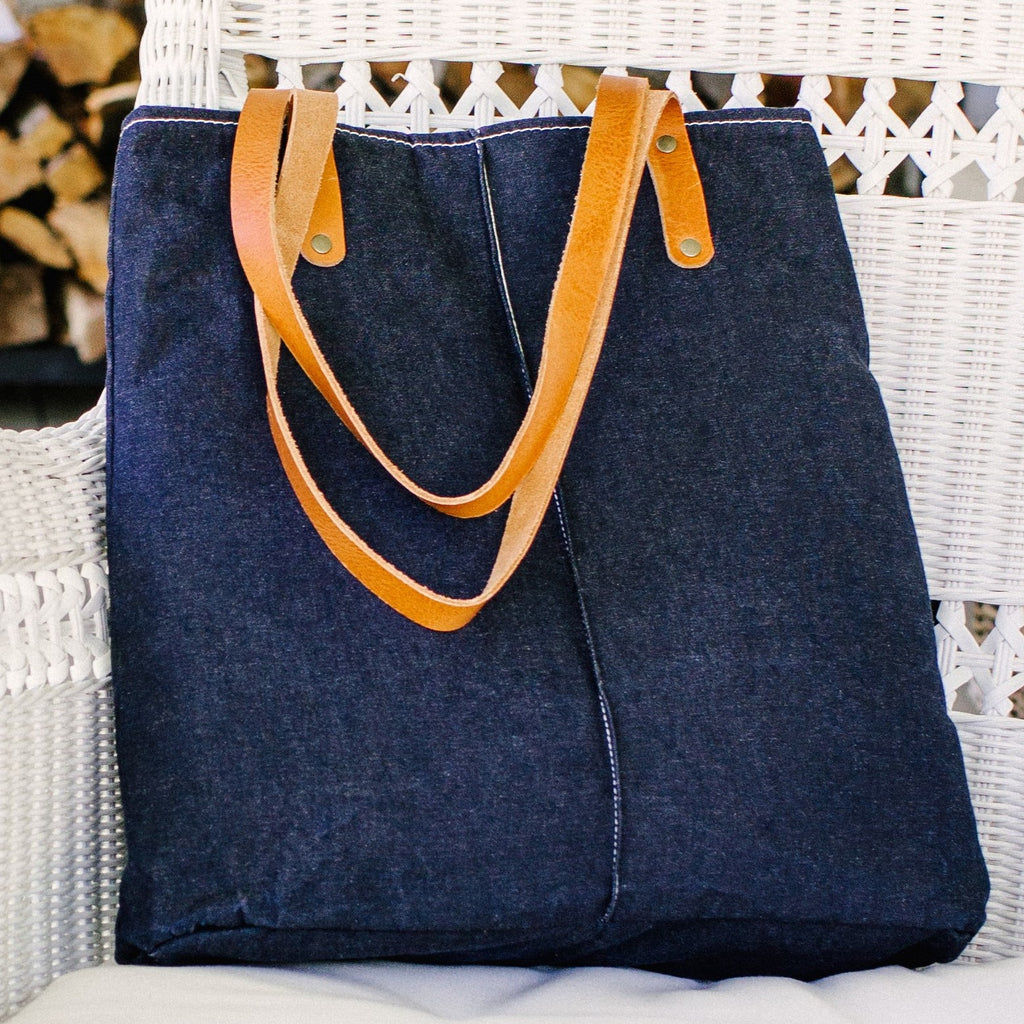 A dark blue waxed denim bag with brown leather straps sits on a white wicker chair