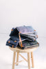 a pile of waxed denim bags and other crossbody bags sits on a wooden stool