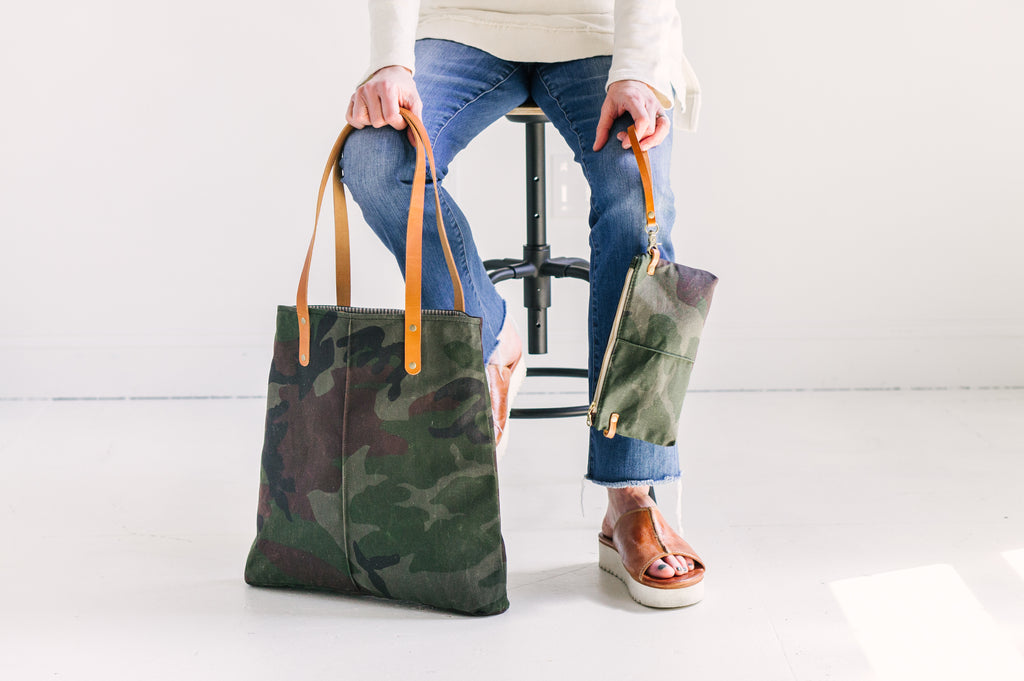 A woman sits on a stool in front of a white wall and holds a camouflage oil cloth tote bag with leather straps