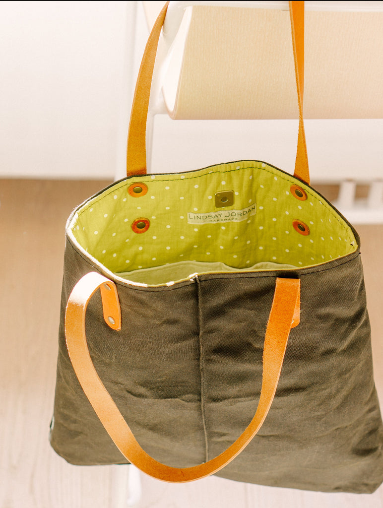 An army green waxed canvas bag with brown leather straps is held open to show bright green polka dot lining fabric