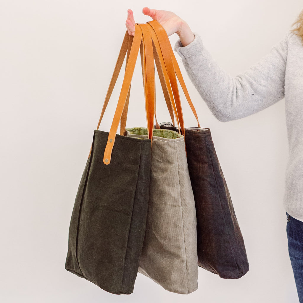 a woman holds a beige waxed corduroy bag with brown leather straps along with two other waxed corduroy bags