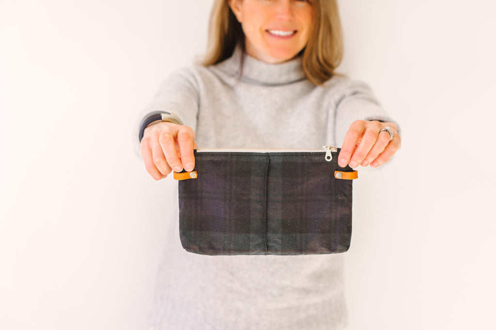 A woman wears aa gray sweater and holds a blackwatch plaid wristlet clutch