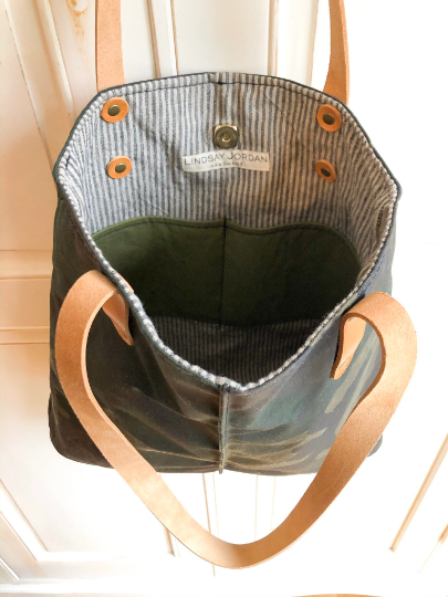A camouflage oil cloth tote bag hangs on a door and is open to showcase inner lining and pockets
