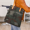 A woman wears jeans and a brown leather jacket and holds a camo oil cloth tote bag with leather straps.