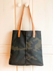 A camouflage oil cloth tote bag with leather straps hangs on a white wooden door