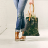 A woman wearing jeans and leather boots stands and holds a camouflage oil cloth tote bag with leather straps