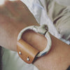 A brown leather and Zamak snaffle bit bracelet is worn on a wrist
