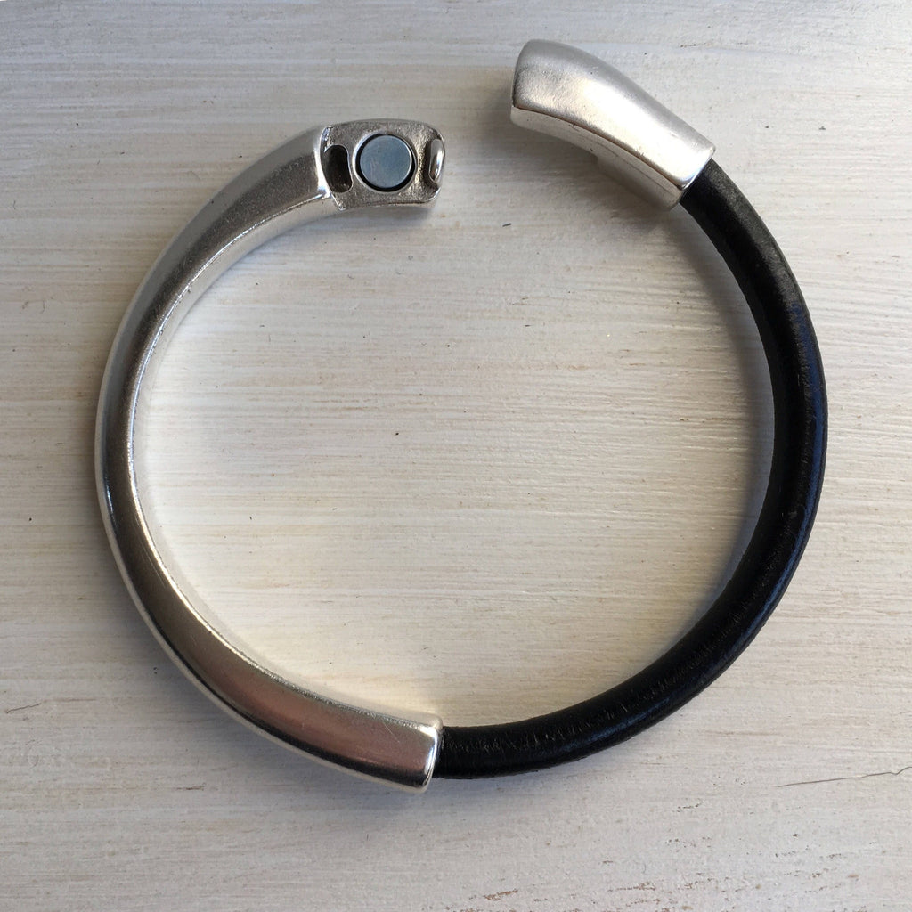 A black leather bracelet with silver clasp sits on a white table with the clasp slightly open