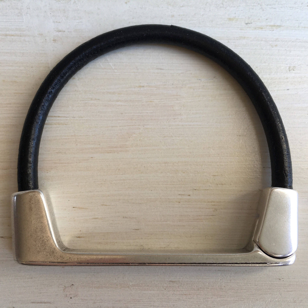 A thin black leather bracelet with silver straight bar clasp sits on a white table