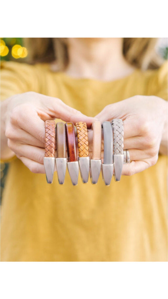 A woman wearing a yellow top holds up a variety of braided leather bracelets with magnetic clasp