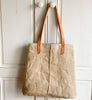 a tan waxed linen bag with brown leather straps hands on a white wooden door