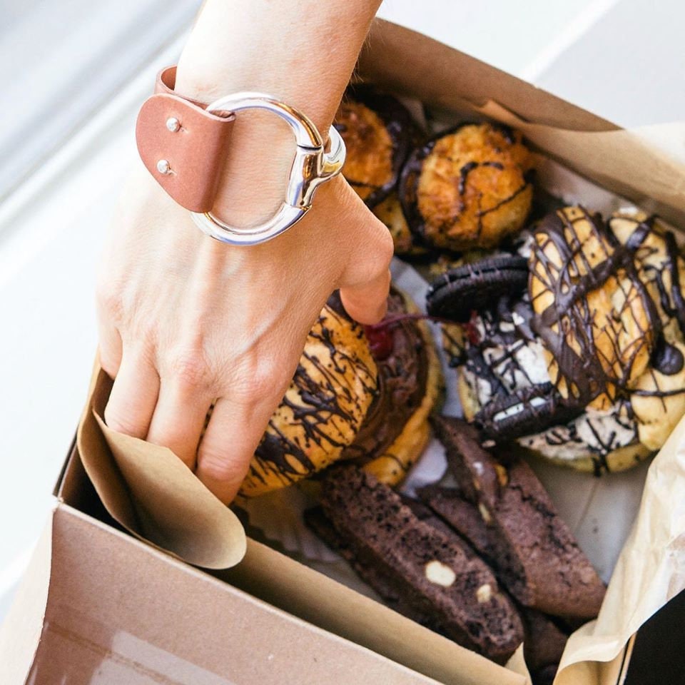 A woman reaches into a box of donuts while wearing a large brown leather and Zamak snaffle bit bracelet