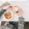 a woman wears a brown leather and dark gray thin leather bracelet while holding a coffee and a croissant