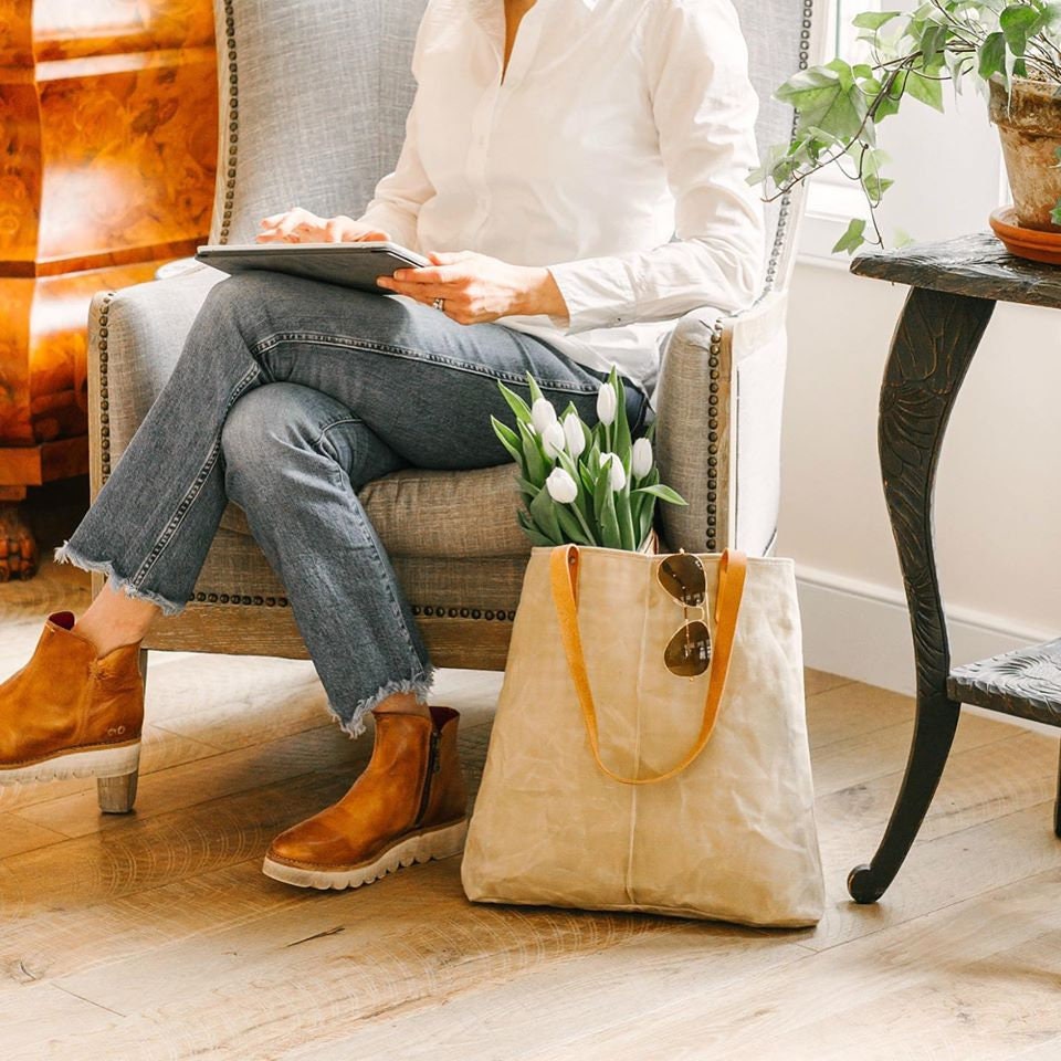 A woman wears jeans and a white shirt and sits on a gray chair with a tan waxed linen bag holding white tulips