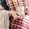 A woman wears a brown leather and Zamak snaffle bit bracelet on her wrist with a plaid shawl and ceramic mug