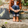 a woman sits on stone steps wearing jeans, leather boots, leather jacket and a brown leather multi strand cuff bracelet