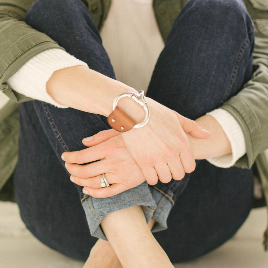 a woman sits cross-legged on the ground wearing jeans, an olive green jacket and a large brown leather and Zamak snaffle bit bracelet