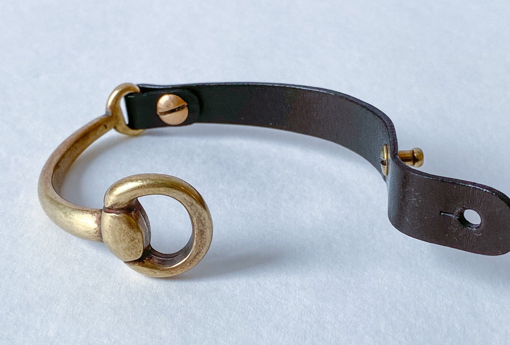 A black leather and antique brass snaffle bit bracelet sits on a white table with the clasp undone