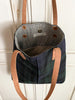 A green and navy blue plaid oil cloth bag with brown leather straps and striiped linen lining hands on a white wooden door