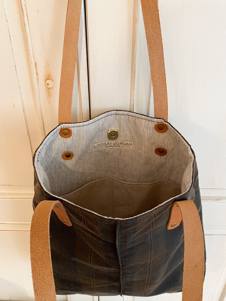 Gold Brown Plaid oil cloth tote with brown leather straps and striped linen lining hangs on a white wooden door