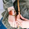 a woman wears a brown leather bracelet and shows off a brown leather strap for crossbody pouch bag