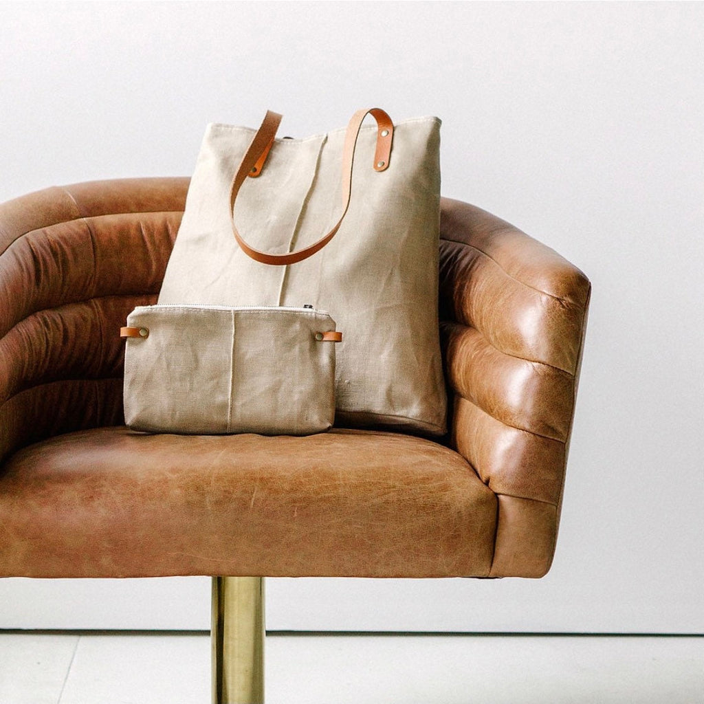a tan waxed linen bag with brown leather straps sits on a brown leather chair